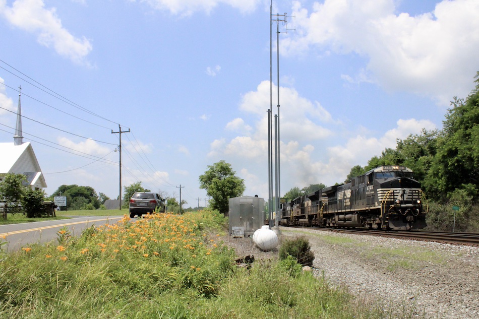 A Norfolk Southern intermodal train passes by orange daylilies and a white church in Virginia.
