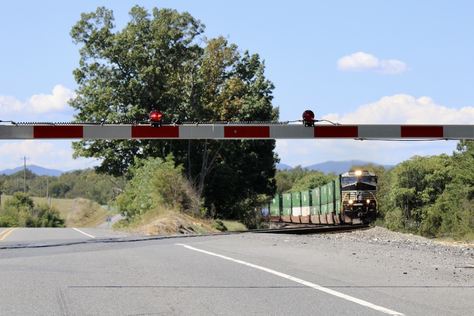 A train approaches a road crossing with the gates down with mountains seen in the distance